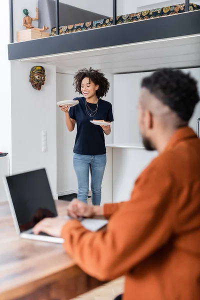 Alegre afroamericana mujer sosteniendo platos con sándwiches cerca borrosa novio usando portátil mientras trabaja desde casa - foto de stock