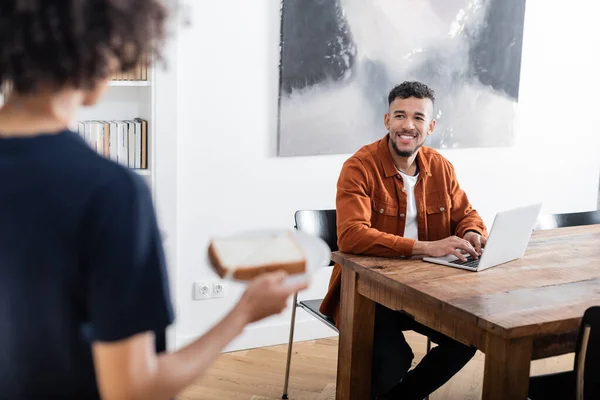 Fröhlicher afrikanisch-amerikanischer Mann mit Laptop und Blick auf verschwommene Freundin, die Teller mit Sandwich hält — Stockfoto