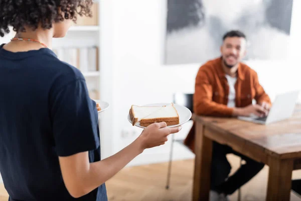 Frisée afro-américaine femme tenant la plaque avec sandwich près petit ami flou en utilisant un ordinateur portable — Photo de stock