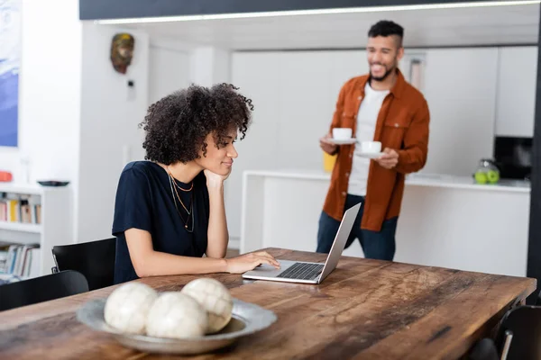 Ricci donna africana americana utilizzando computer portatile vicino fidanzato felice in possesso di tazze di caffè — Foto stock