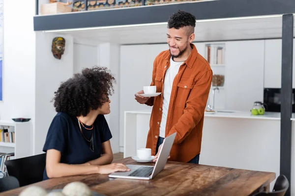 Pleased african american woman using laptop near happy boyfriend holding cups of coffee — Stock Photo