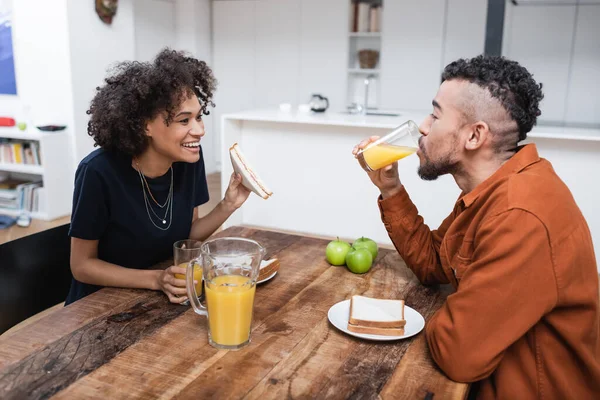 Happy african american woman holding sandwich while boyfriend drinking orange juice — Stock Photo