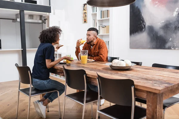 Feliz africana americana mujer celebración sándwich mientras novio beber jugo de naranja durante almuerzo - foto de stock