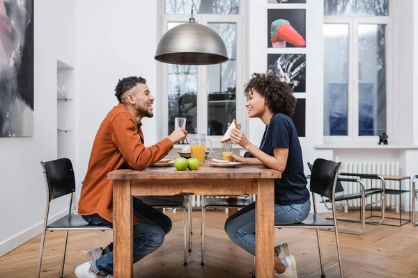 Felice donna afro-americana che tiene il panino e pranza con il fidanzato allegro — Foto stock