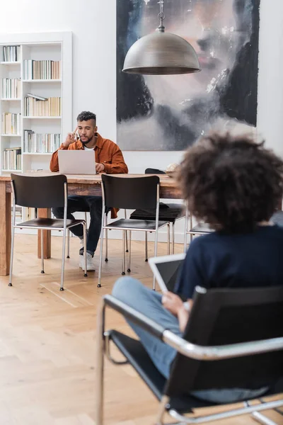 Freelance afroamericano che parla su smartphone che lavora da casa vicino alla fidanzata riccia usando tablet digitale — Foto stock