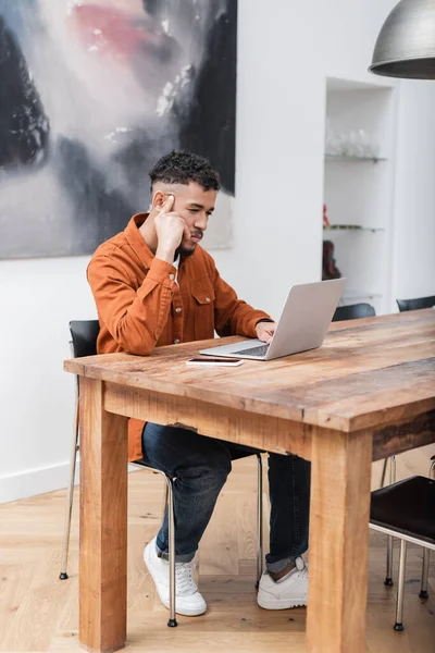 Pensativo afroamericano freelancer utilizando el ordenador portátil mientras trabaja desde casa - foto de stock