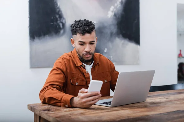 Freelancer afroamericano sosteniendo teléfono inteligente cerca de la computadora portátil mientras trabaja desde casa - foto de stock