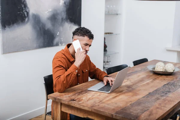 Freelancer afro-americano falando no smartphone perto do laptop enquanto trabalha em casa — Fotografia de Stock
