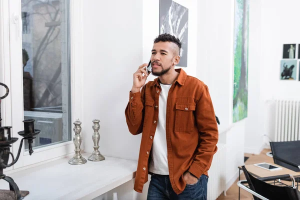 Lockiger afrikanisch-amerikanischer Mann telefoniert, während er mit der Hand in der Tasche in einer modernen Wohnung steht — Stockfoto