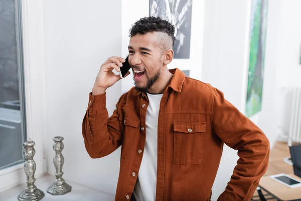 Hombre afroamericano emocionado hablando por teléfono móvil en apartamento moderno - foto de stock