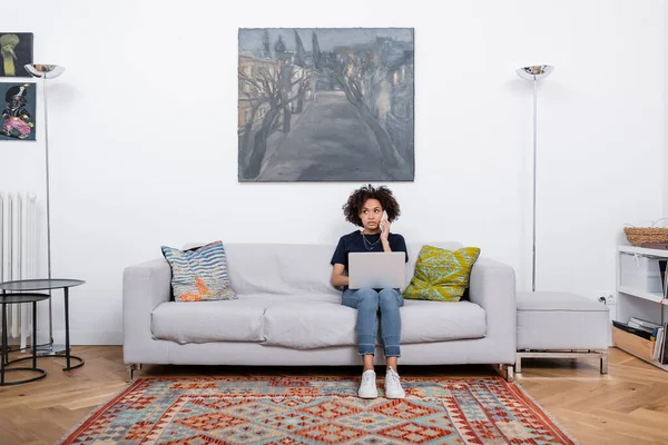 Curly african american woman sitting on couch and talking on smartphone near laptop — Stock Photo