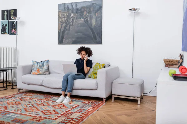 Giovane donna africana americana seduta sul divano e che parla sul telefono cellulare vicino al computer portatile — Foto stock