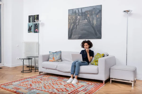 Mujer afroamericana joven sentada en el sofá y grabando el mensaje de voz en el teléfono móvil mientras mira el portátil - foto de stock