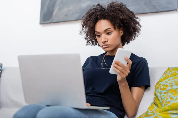 Lockige Afroamerikanerin hält Handy in der Hand, während sie auf Laptop schaut — Stockfoto