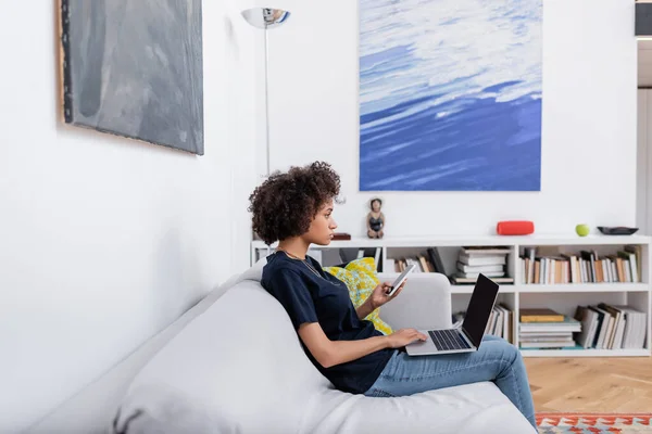 Vista lateral de la mujer afroamericana rizada sosteniendo el teléfono móvil cerca del ordenador portátil mientras está sentado en el sofá — Stock Photo