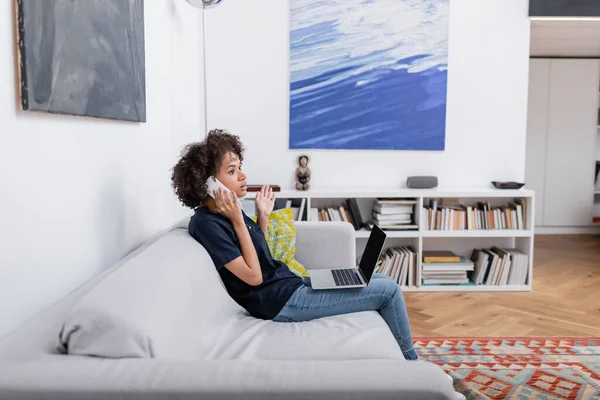 Удивленный африканский американец женщина разговаривает на мобильном телефоне рядом с ноутбуком, сидя на диване — стоковое фото