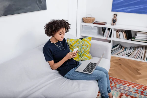 Curly african american woman using cellphone near laptop while sitting on sofa — Stock Photo