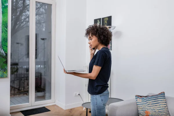 Mujer afroamericana rizada hablando en el teléfono inteligente y sosteniendo el ordenador portátil en la sala de estar - foto de stock