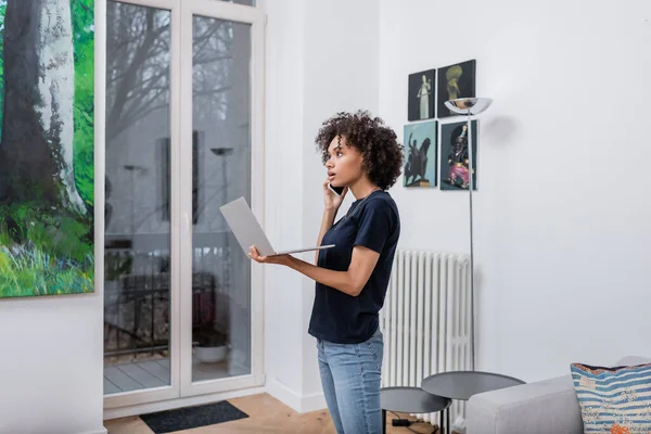 Junge Afroamerikanerin spricht auf Smartphone und hält Laptop im Wohnzimmer — Stockfoto