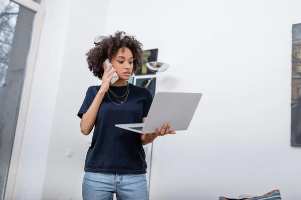 Encaracolado afro-americano mulher falando no telefone celular e segurando laptop no apartamento moderno — Fotografia de Stock