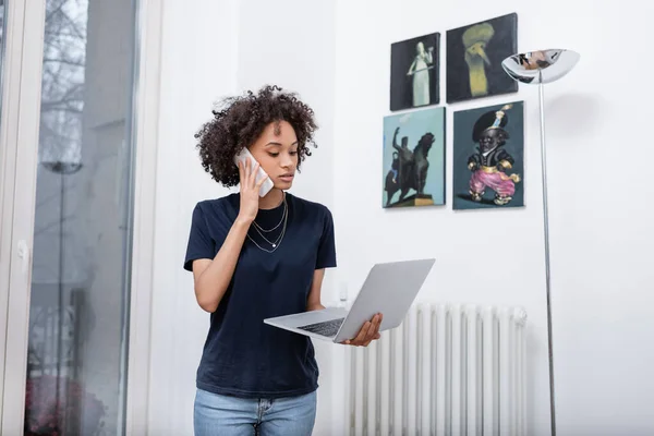 Молодая африканская американка разговаривает по мобильному телефону и держит ноутбук в современной квартире — стоковое фото