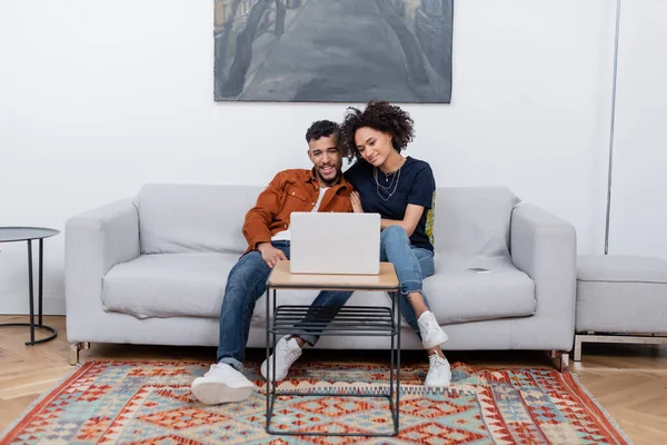 Молода афроамериканська пара посміхається, дивлячись кіно на ноутбуку в сучасній квартирі. — стокове фото