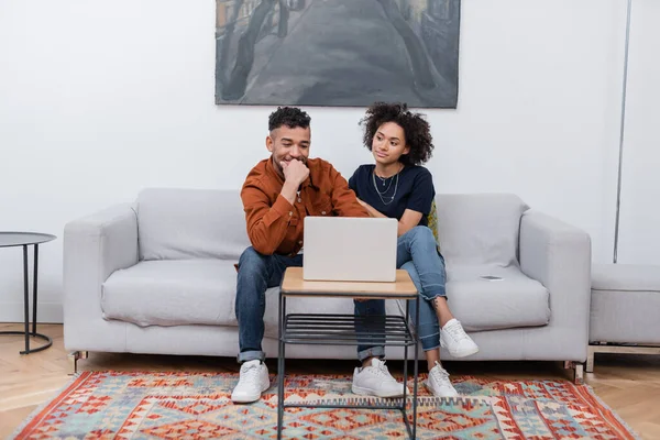 Heureux jeune couple afro-américain souriant tout en regardant un film sur ordinateur portable dans un appartement moderne — Photo de stock
