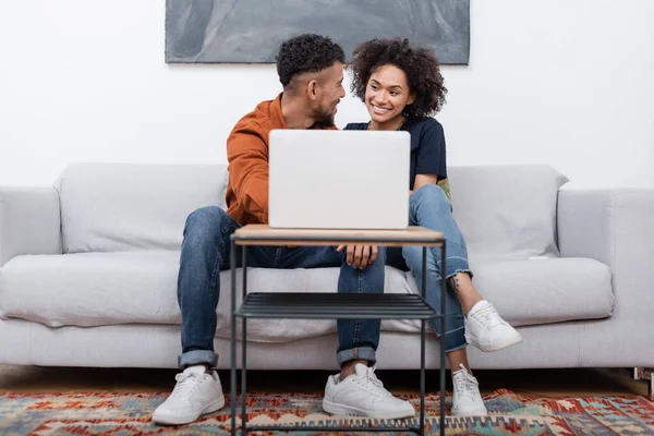 Feliz jovem casal afro-americano sorrindo enquanto olham um para o outro perto do laptop no apartamento moderno — Fotografia de Stock