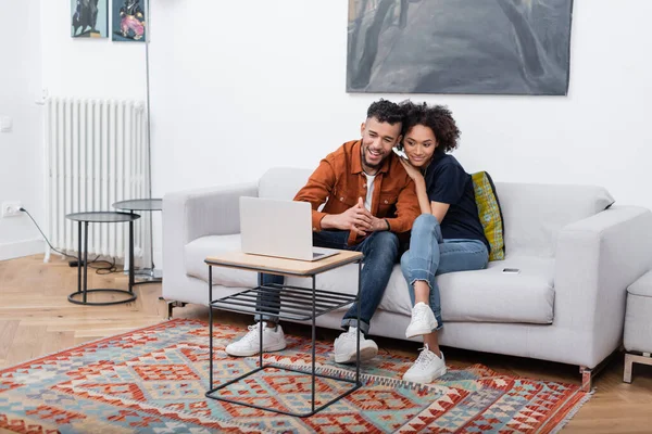 Alegre jovem casal afro-americano sorrindo enquanto assiste filme no laptop no apartamento moderno — Fotografia de Stock
