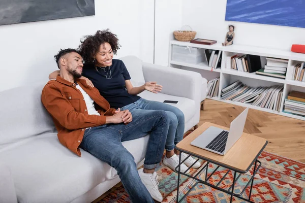 Alegre joven afroamericana mujer apuntando con la mano a la computadora portátil mientras ve la película con el novio rizado - foto de stock