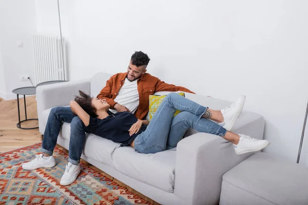 Весела молода афроамериканська пара, яка дивиться один на одного, відпочиваючи на дивані в сучасній квартирі. — стокове фото