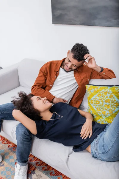 Heureux jeune couple afro-américain regardant l'autre tout en se reposant sur le canapé dans un appartement moderne — Photo de stock