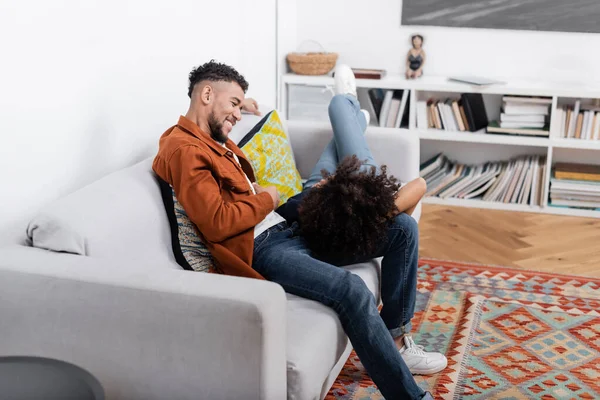 Fröhlicher junger afrikanisch-amerikanischer Mann schaut Freundin an, die auf Couch in moderner Wohnung ruht — Stockfoto