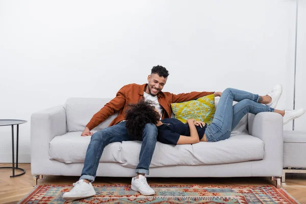 Heureux homme afro-américain regardant petite amie reposant sur le canapé dans un appartement moderne — Photo de stock