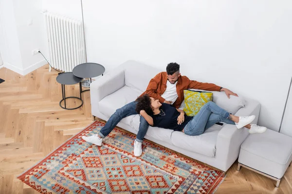 Vista de ángulo alto de la alegre pareja afroamericana mirándose mientras descansan en el sofá en el apartamento moderno - foto de stock