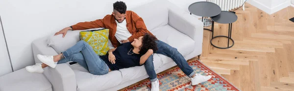 Vue grand angle du couple afro-américain joyeux se regardant tout en se reposant sur le canapé dans un appartement moderne, bannière — Photo de stock