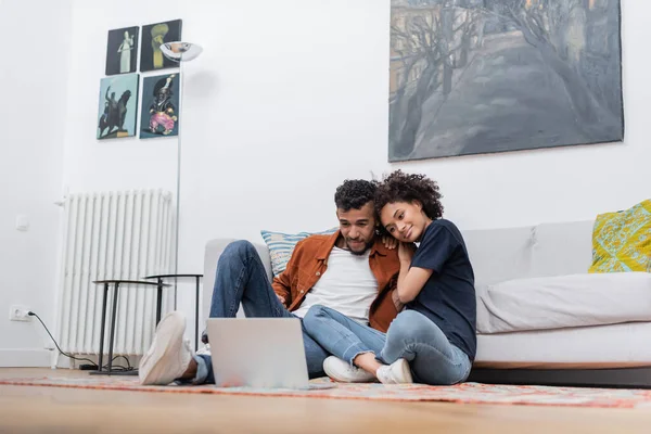 Alegre africano americano pareja sentado en alfombra y viendo película en laptop - foto de stock