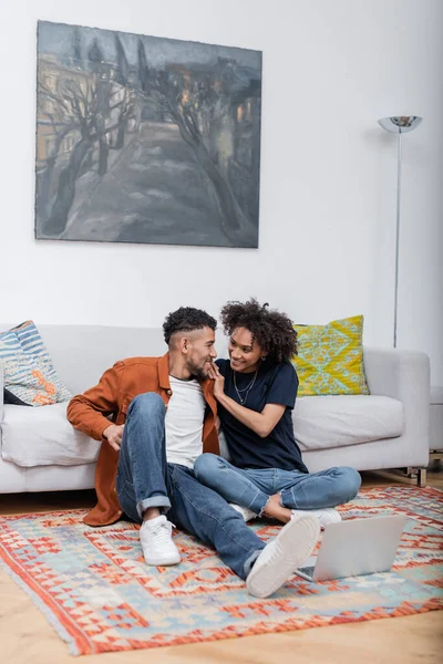 Веселая африканская американская пара сидит на ковре и смотрит друг на друга рядом с ноутбуком — стоковое фото
