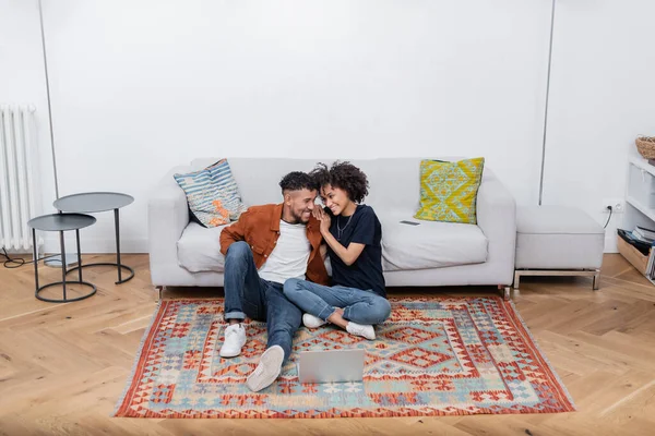 Fröhliches afrikanisch-amerikanisches Paar auf Teppich sitzend und sich in der Nähe von Laptop und Smartphone auf dem Sofa anschauend — Stockfoto