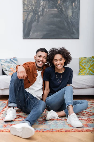 Joyeuse femme afro-américaine avec tatouage assis près heureux petit ami sur le tapis dans un appartement moderne — Photo de stock