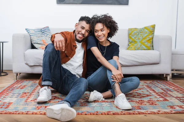 Alegre africana americana mujer con tatuaje sentado cerca feliz novio en alfombra en moderno apartamento - foto de stock