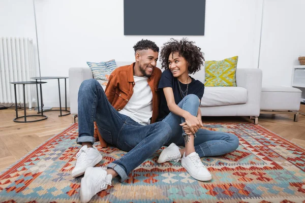 Joyful african american woman with tattoo and happy man looking at each other while sitting on carpet in modern apartment — Stock Photo