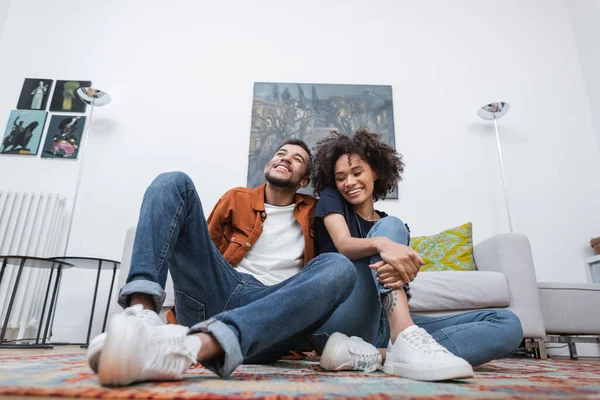 Low angle view of positive african american woman with tattoo sitting near happy boyfriend on carpet — Stock Photo