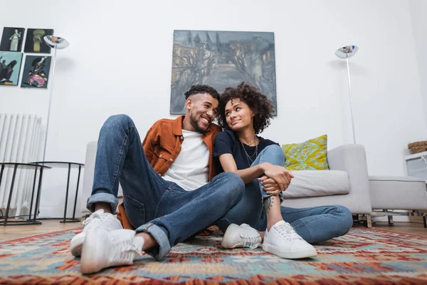 Vista a basso angolo di gioiosa donna africana americana con tatuaggio e uomo felice che si guarda mentre si siede sul tappeto — Foto stock