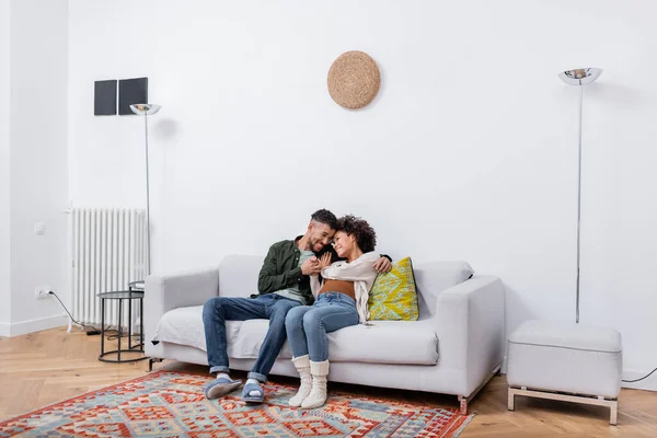 Embarazada africana americana mujer sentado con complacido marido en sofá en moderno apartamento - foto de stock