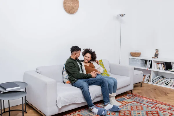 Mujer afroamericana feliz y embarazada sentada con marido feliz en el sofá - foto de stock