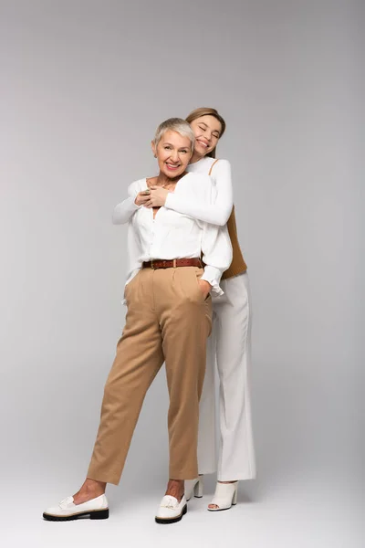 Longitud completa de la joven hija feliz abrazando a la madre de mediana edad de pie con las manos en los bolsillos en gris — Stock Photo