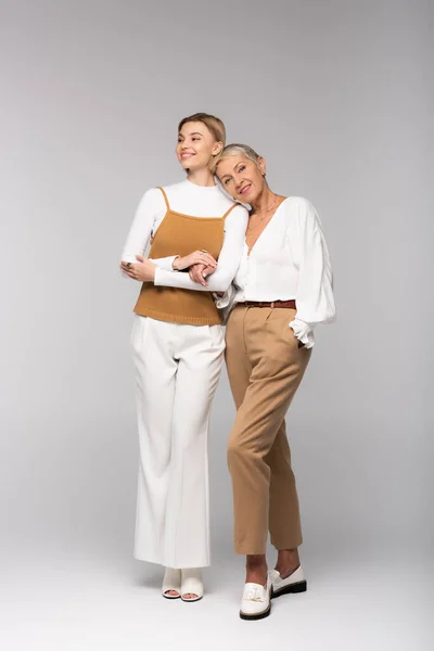 Full length of cheerful middle aged mother standing with hand in pocket near young daughter on grey — Stock Photo