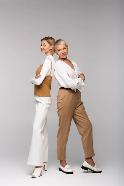 Full length of cheerful middle aged mother standing with crossed arms near young stylish daughter on grey — Stock Photo