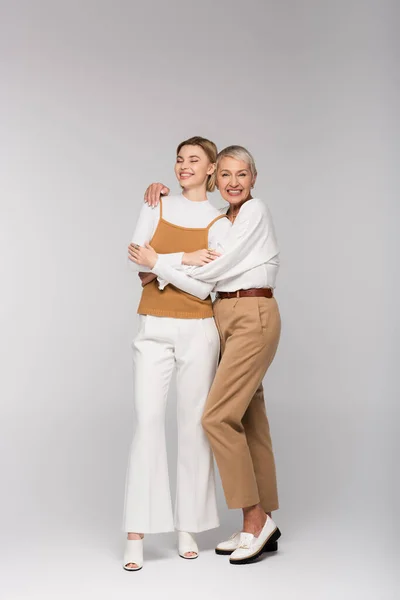 Longitud completa de la madre de mediana edad positiva abrazando a la hija joven en gris - foto de stock